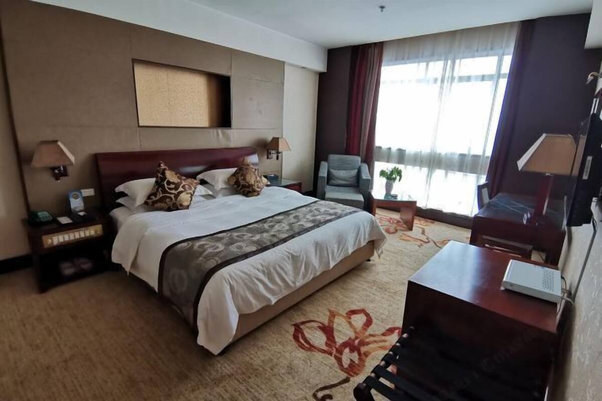 Une chambre d’hôtel avec un grand lit, deux tables de chevet, une chaise, un bureau, une télévision et une fenêtre avec des rideaux.