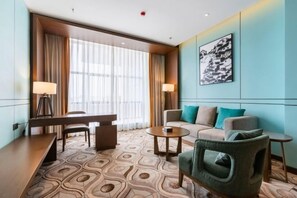 Habitación de hotel con una mesa de comedor, un sofá, una silla, una mesita de centro y un amplio ventanal con cortinas.
