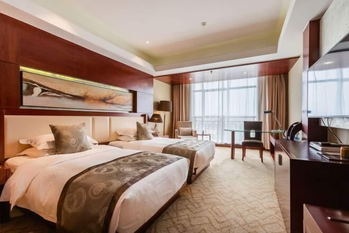 Une chambre d’hôtel avec deux lits, un bureau, une chaise, une télévision et une grande fenêtre avec des rideaux.