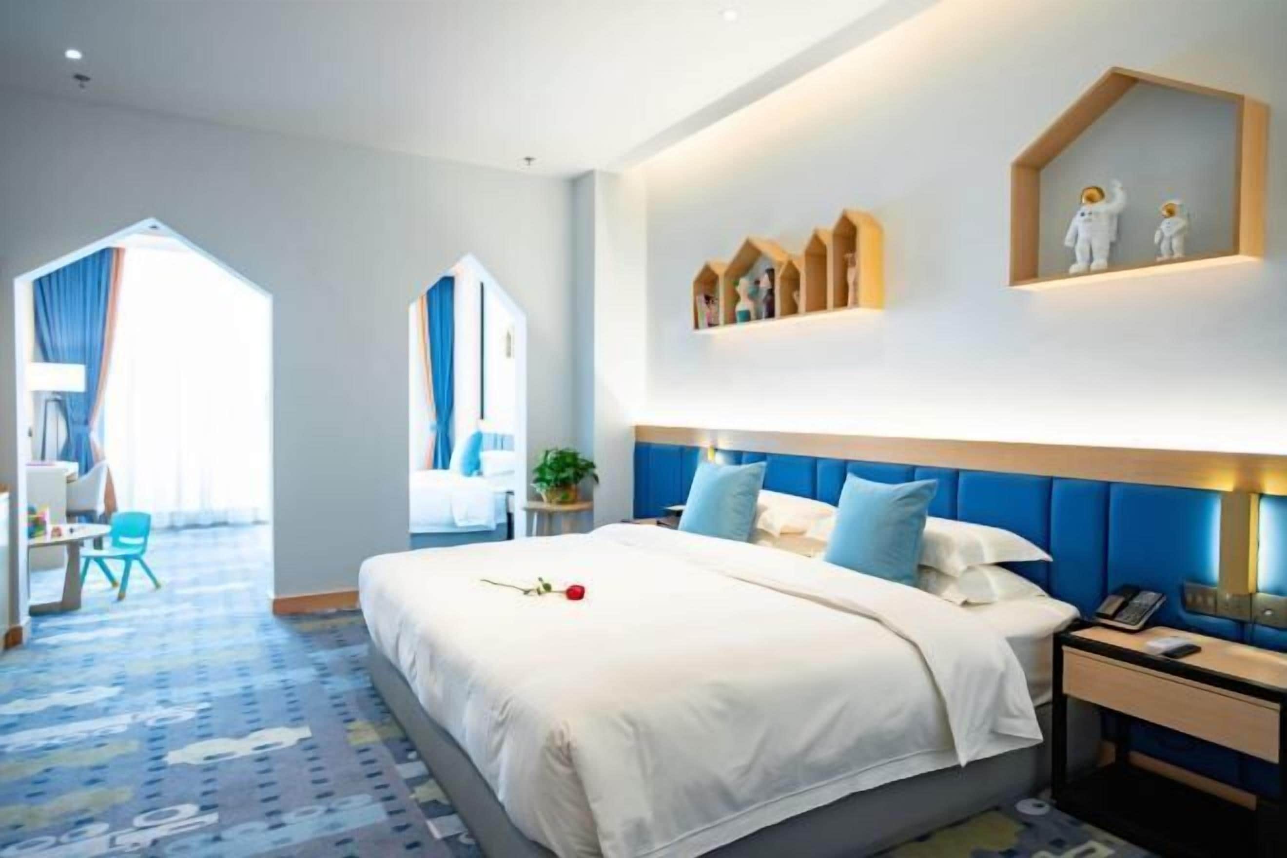Habitación de hotel con una cama grande, un escritorio y una silla. Hay una ventana con cortinas, una planta en el escritorio y un estante con objetos decorativos en la pared.