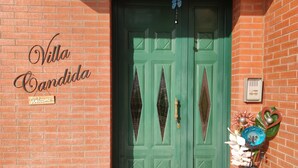 Puertas verdes dobles con ventanas en forma de diamante, un cartel de bienvenida y un jarrón decorativo con flores.