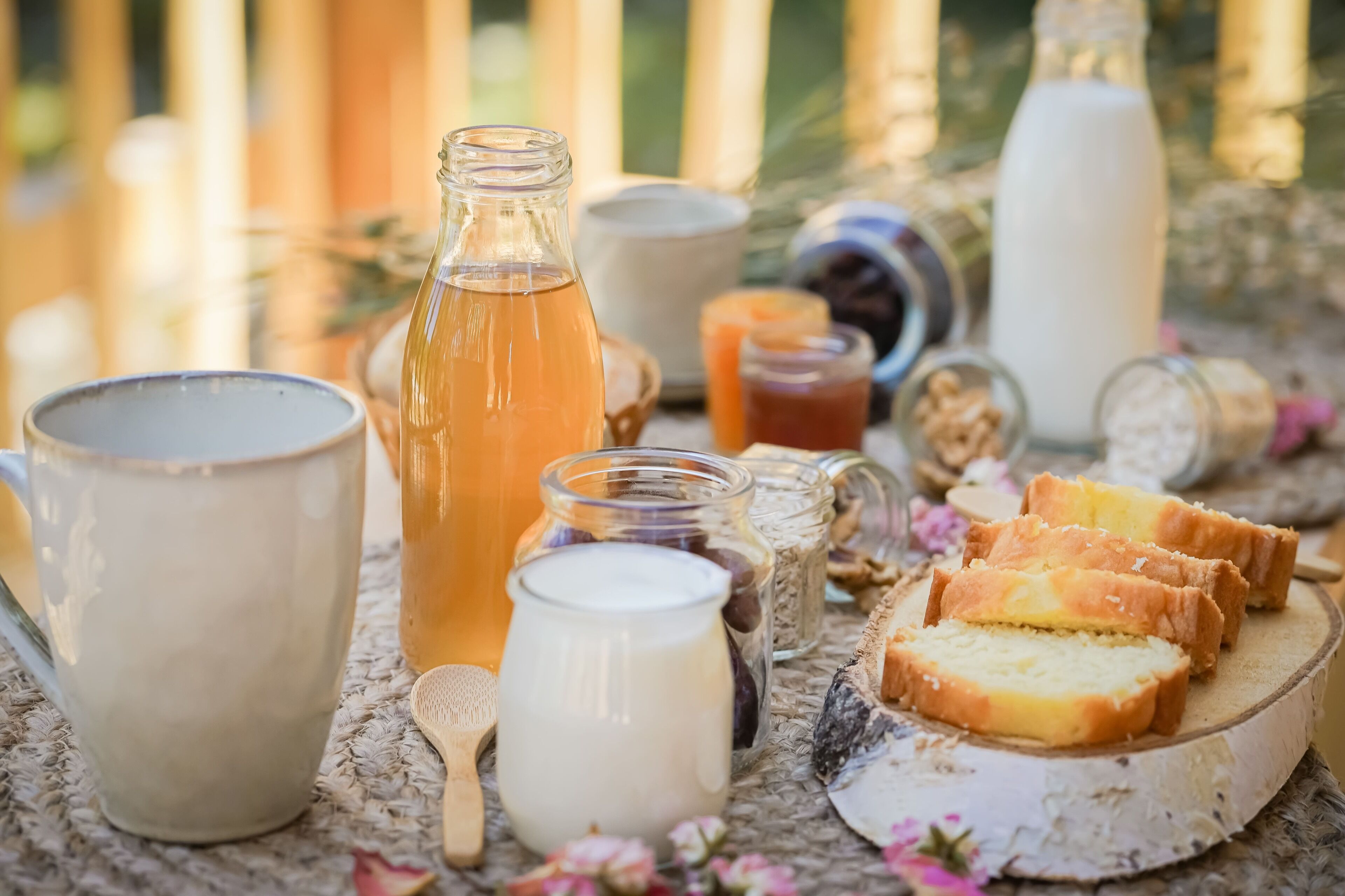 A rustic outdoor setting with a table displaying various food items, including bread, jars of preserves, and a milk jug.