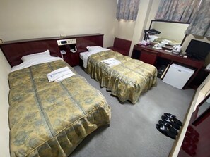 Habitación de hotel con dos camas, un escritorio con computadora, un refrigerador pequeño y un par de pantuflas en el suelo.