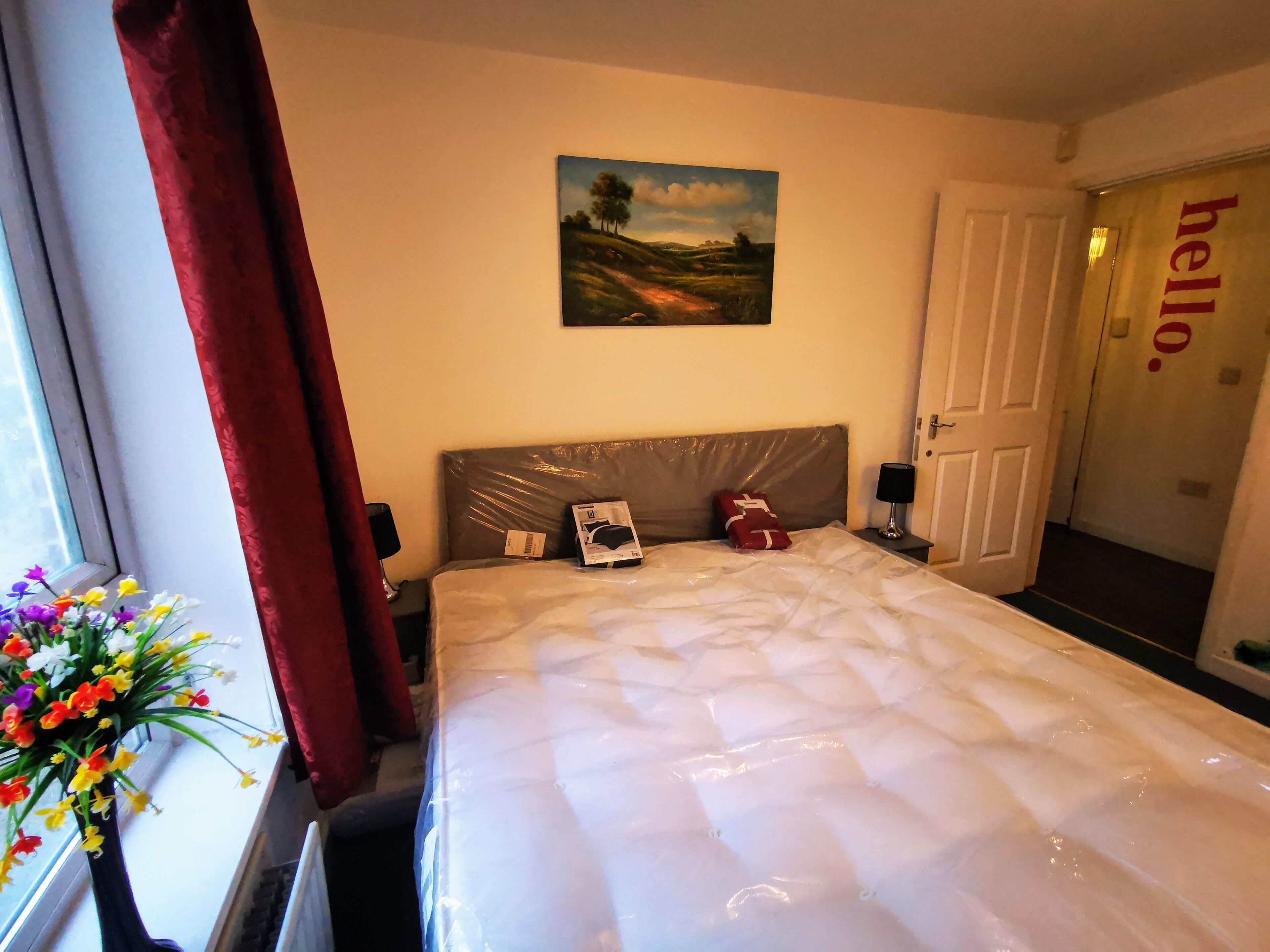 A hotel room with a bed, a nightstand, a lamp, a painting on the wall, a window with curtains, and a door with a 'hello' sign.
