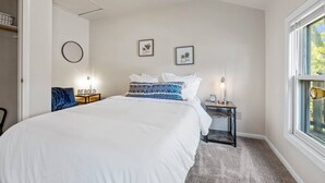 A bedroom with a large bed, a bedside table with a lamp, a clock, and a window with a view of greenery. - Sunny, Furnished  2 Bedroom Upper Apt Near Downtown Green Bay (Green Bay)