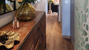 A hallway with a wooden floor, a mirror on the left, a dresser with a vase of flowers, and a patterned wall on the right.