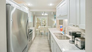 Una cocina moderna con armarios blancos, electrodomésticos de acero inoxidable, un comedor con mesa y sillas, y vistas a través de un ventanal.