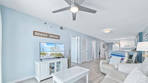 Uma sala de estar com televisão, móveis brancos, um ventilador de teto e uma placa com a inscrição "BEACH HOUSE".