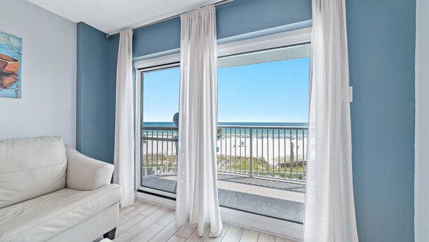 Quarto com sofá bege, uma janela grande com cortinas brancas e vista para a praia.