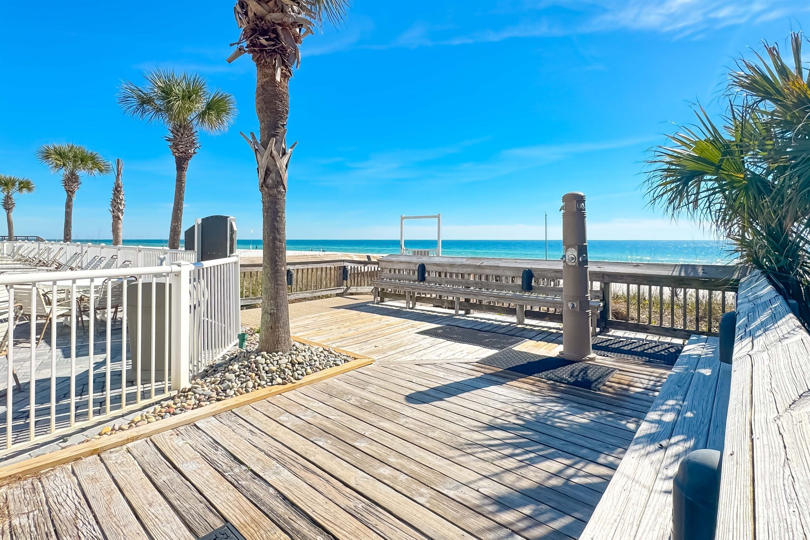 A wooden boardwalk with benches and palm trees, leading to a beach with clear blue water.