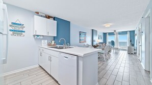 Cozinha moderna com armários brancos, lava-louças e pia. Possui área de jantar com mesa e cadeiras, e vista para o exterior através de grandes janelas.