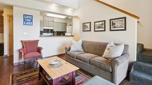 A living room with a sofa, armchair, and a coffee table. There is a kitchen area with a refrigerator and a sink.