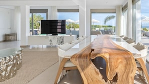 Una sala de estar moderna con una gran mesa de centro de madera, un televisor de pantalla plana y una zona de comedor con mobiliario blanco.