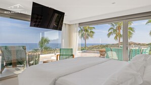 Un dormitorio con una cama grande, un televisor montado en la pared y un balcón con vistas al mar y palmeras.