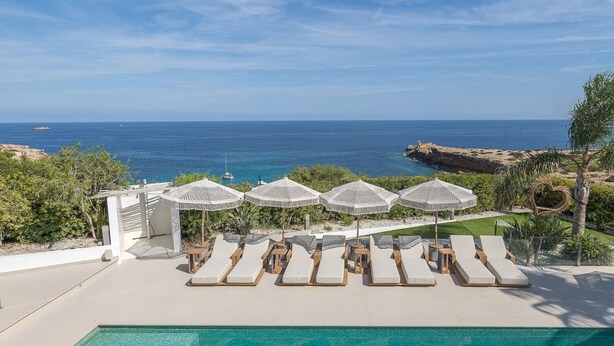 Zona junto a la piscina con sillones de descanso y sombrillas, con vistas al mar azul y transparente.