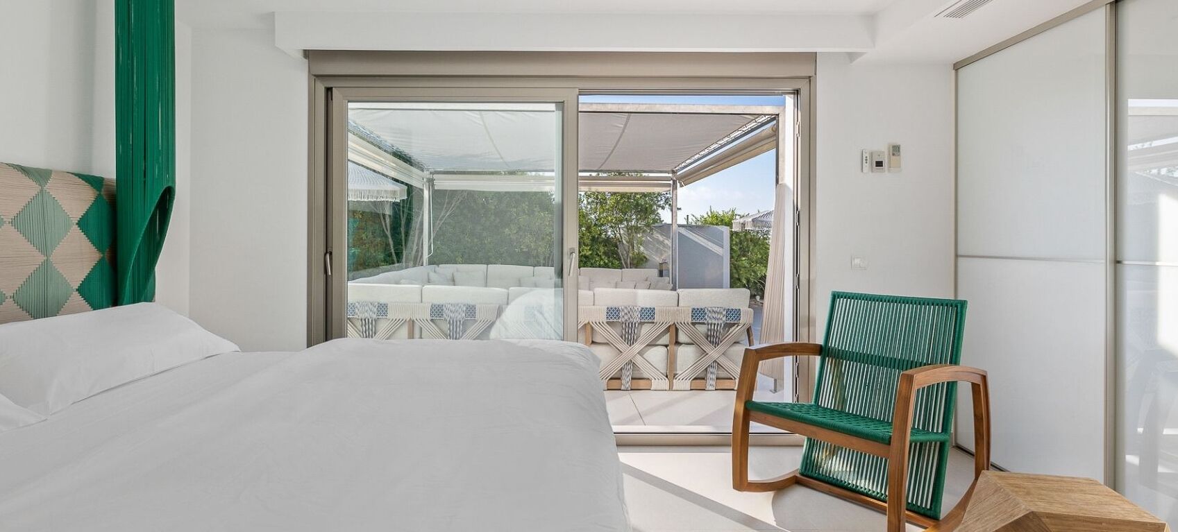 Une chambre avec un lit, un fauteuil vert et une porte-fenêtre coulissante donnant sur un espace extérieur aménagé avec un canapé blanc et des coussins à motifs.