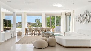 Una sala moderna con un sofá blanco, un comedor con mesa de vidrio y grandes ventanales con vistas al exterior.