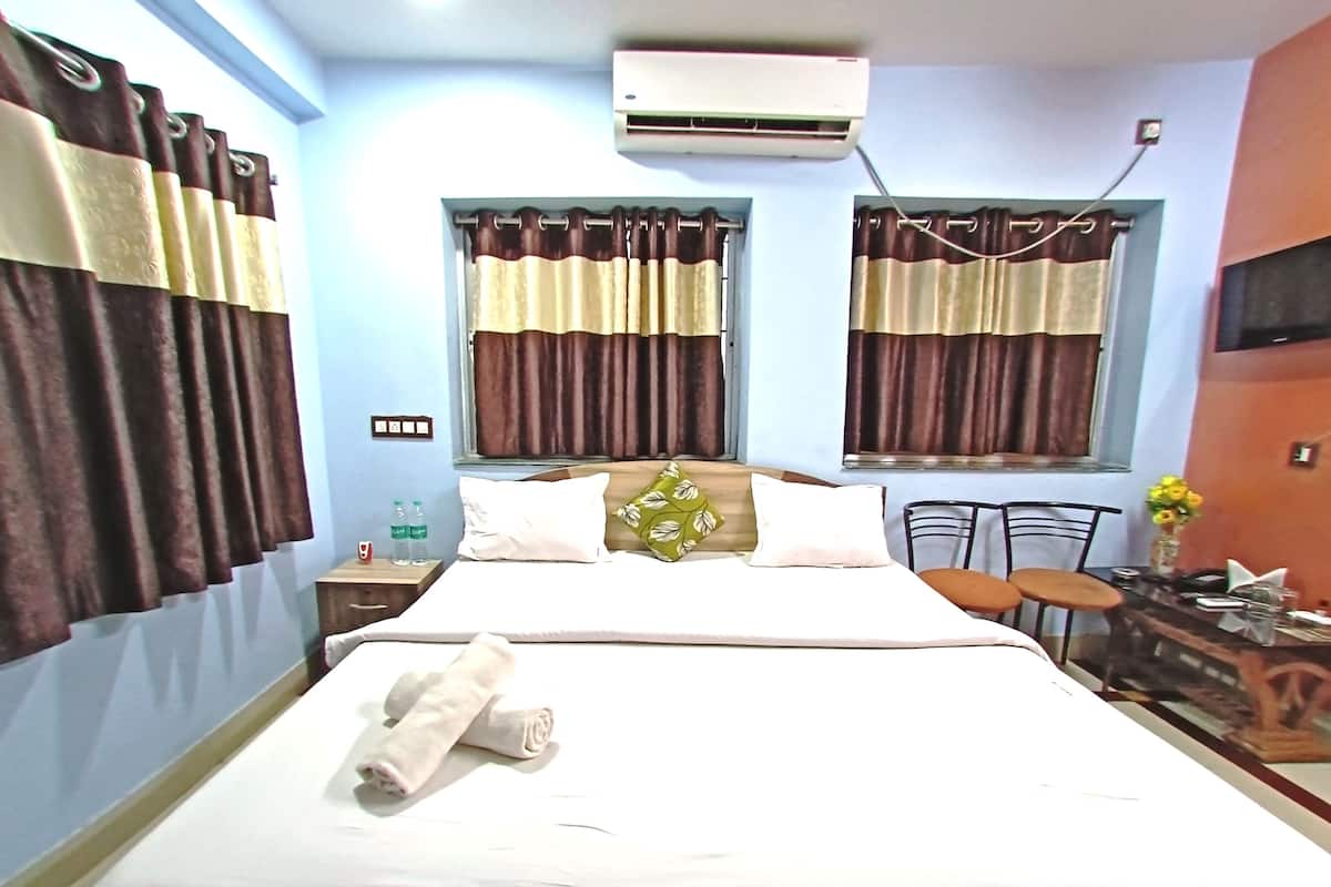 A hotel room with a bed, two windows with curtains, an air conditioning unit, a small table with a chair, and a vase with flowers.