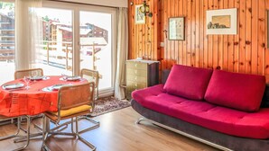 A cozy room with a wooden wall, a red sofa, a dining table set for four, and a view of buildings outside. - Olympic F8 by Interhome (Nendaz)