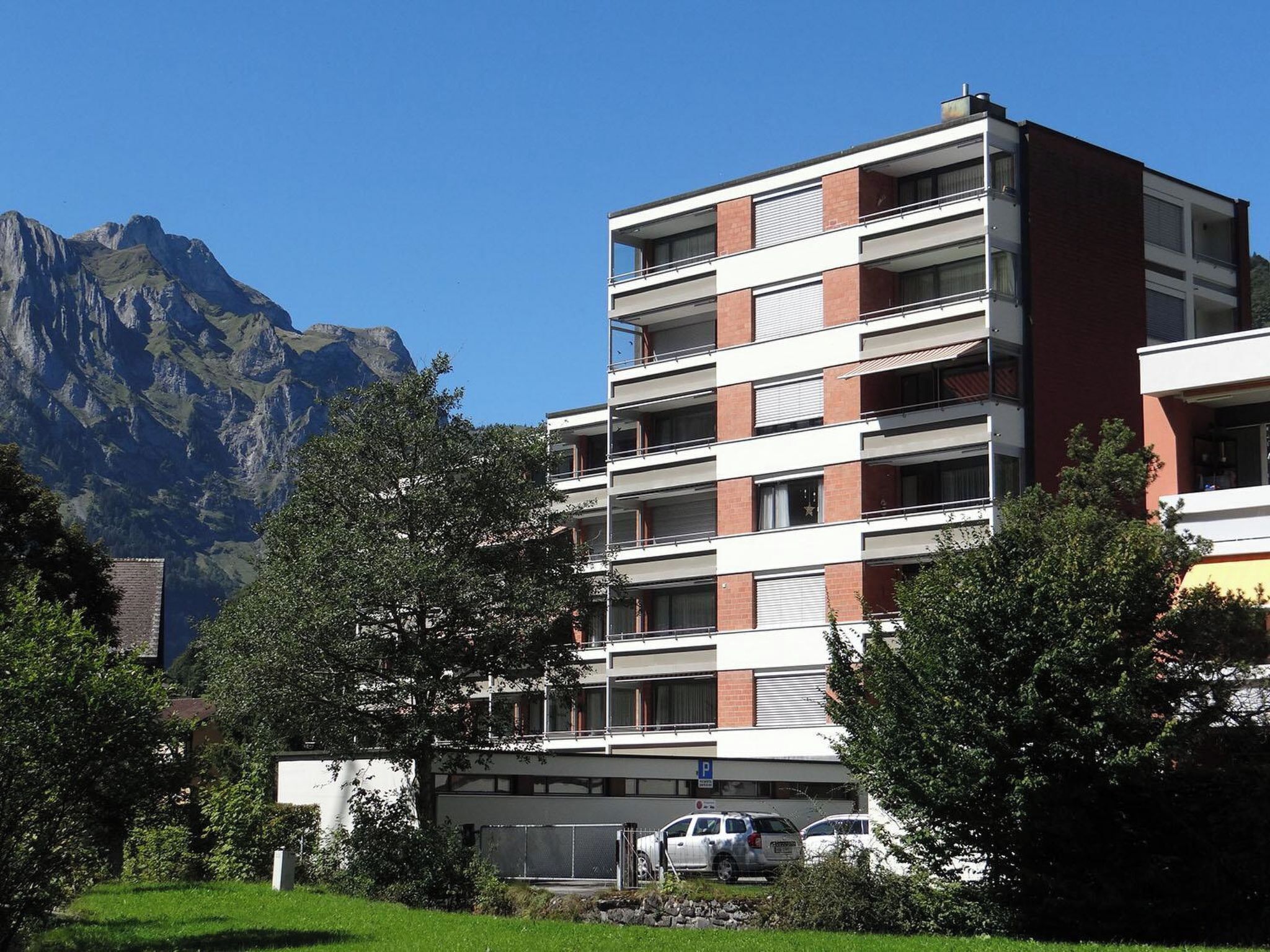 A multi-story building with a flat roof, surrounded by greenery and mountains in the background.
