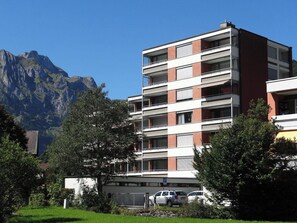 A multi-story building with a flat roof, surrounded by greenery and mountains in the background.