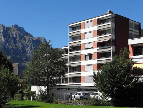 Un edificio de varios pisos con techo plano, rodeado de vegetación y montañas al fondo.