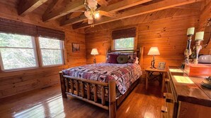 A wooden cabin bedroom with a bed, two windows, a ceiling fan, and a nightstand with a lamp. - Grumpy Bear Cabin-Hot Tub-Pet Friendly (Murphy)