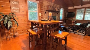A cozy cabin interior with a wooden dining table, four stools, a stone fireplace, and a large window. - Grumpy Bear Cabin-Hot Tub-Pet Friendly (Murphy)