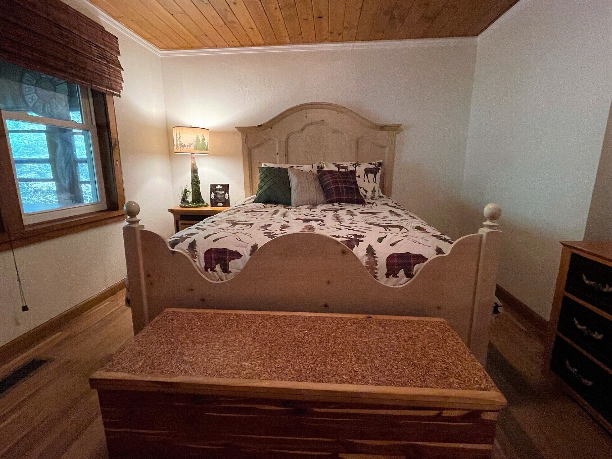 A bedroom with a wooden headboard, a bed with a bear-themed bedspread, a nightstand with a lamp, and a window with blinds.