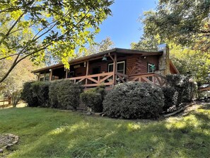 A rustic cabin with a wooden deck, surrounded by well-maintained hedges and a grassy yard.
