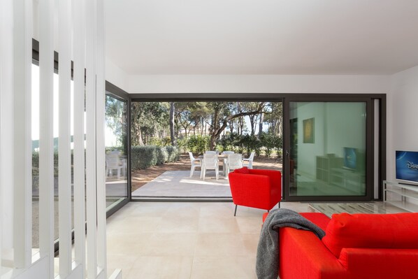 A modern living room with a red sofa, a TV, and a glass door leading to an outdoor patio with white chairs and a table.