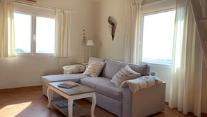 En stue med en grå sofa, et lille hvidt bord og en lampe. To vinduer med gardiner, og en synlig træbjælke i loftet.