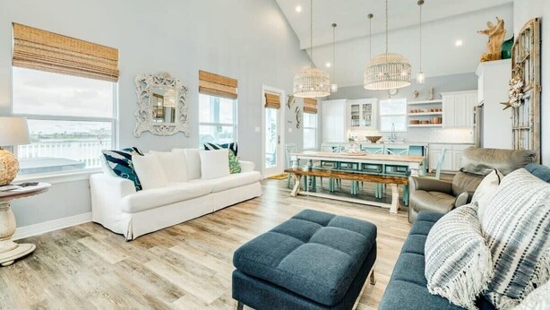 A spacious living room with a white sofa, a blue ottoman, and a grey couch. There is a dining area with a wooden table and chairs, and a kitchen in the background. - Sweet & Salty - Hot Tub, Across from Jetty, Best Ship Watching! (Surfside Beach)