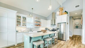 A modern kitchen with white cabinets, a central island with stools, stainless steel appliances, and a light blue accent wall. - Sweet & Salty - Hot Tub, Across from Jetty, Best Ship Watching! (Surfside Beach)