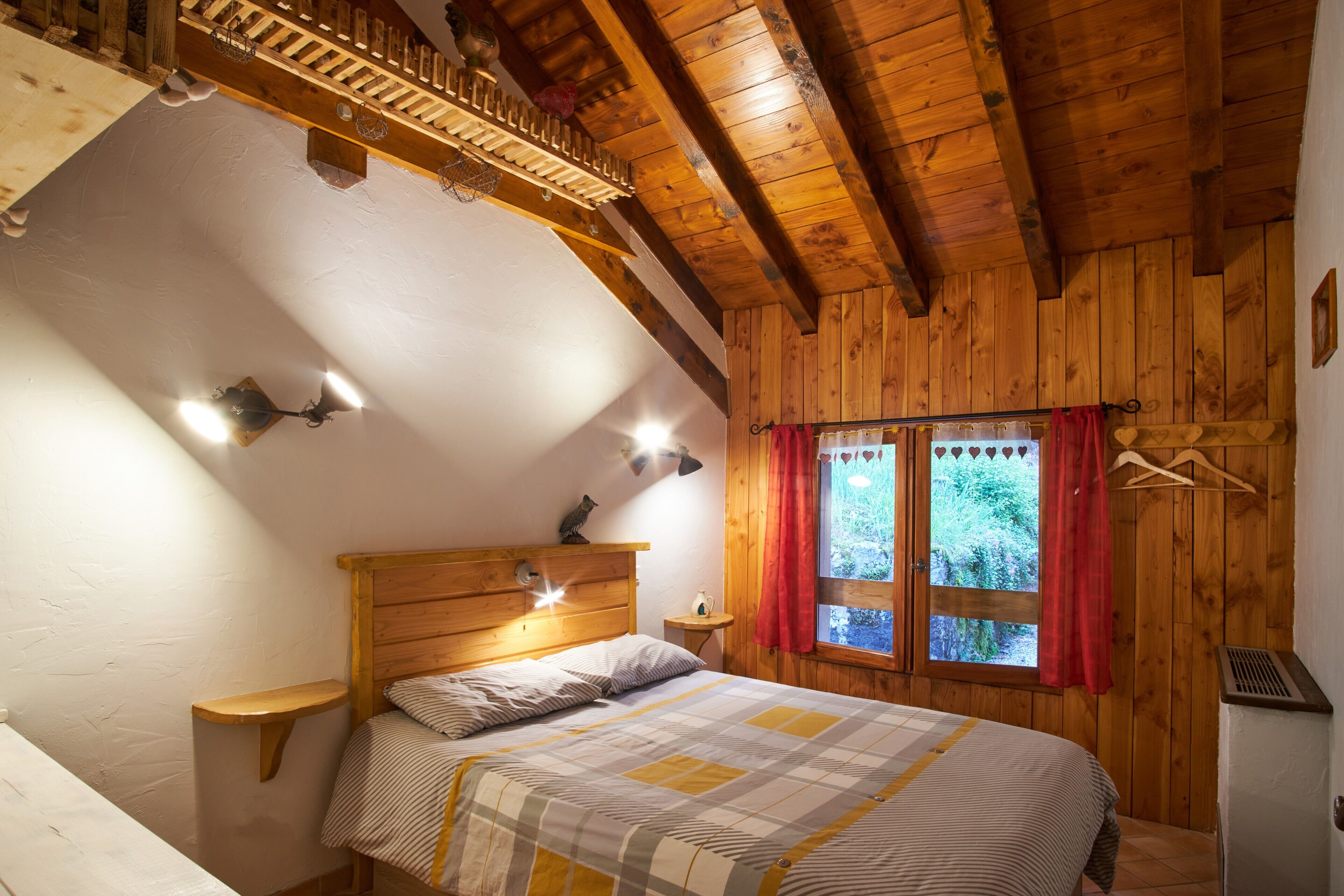 Une chambre avec un plafond en bois, un lit recouvert d’une courtepointe à rayures, une table de chevet, une étagère fixée au mur et une fenêtre avec un rideau rouge.