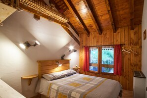 A bedroom with a wooden ceiling, a bed with a striped bedspread, a nightstand, a wall-mounted shelf, and a window with a red curtain. - Holiday Accommodation "Maison de pierre" with beautiful view (Saint-Gervais)