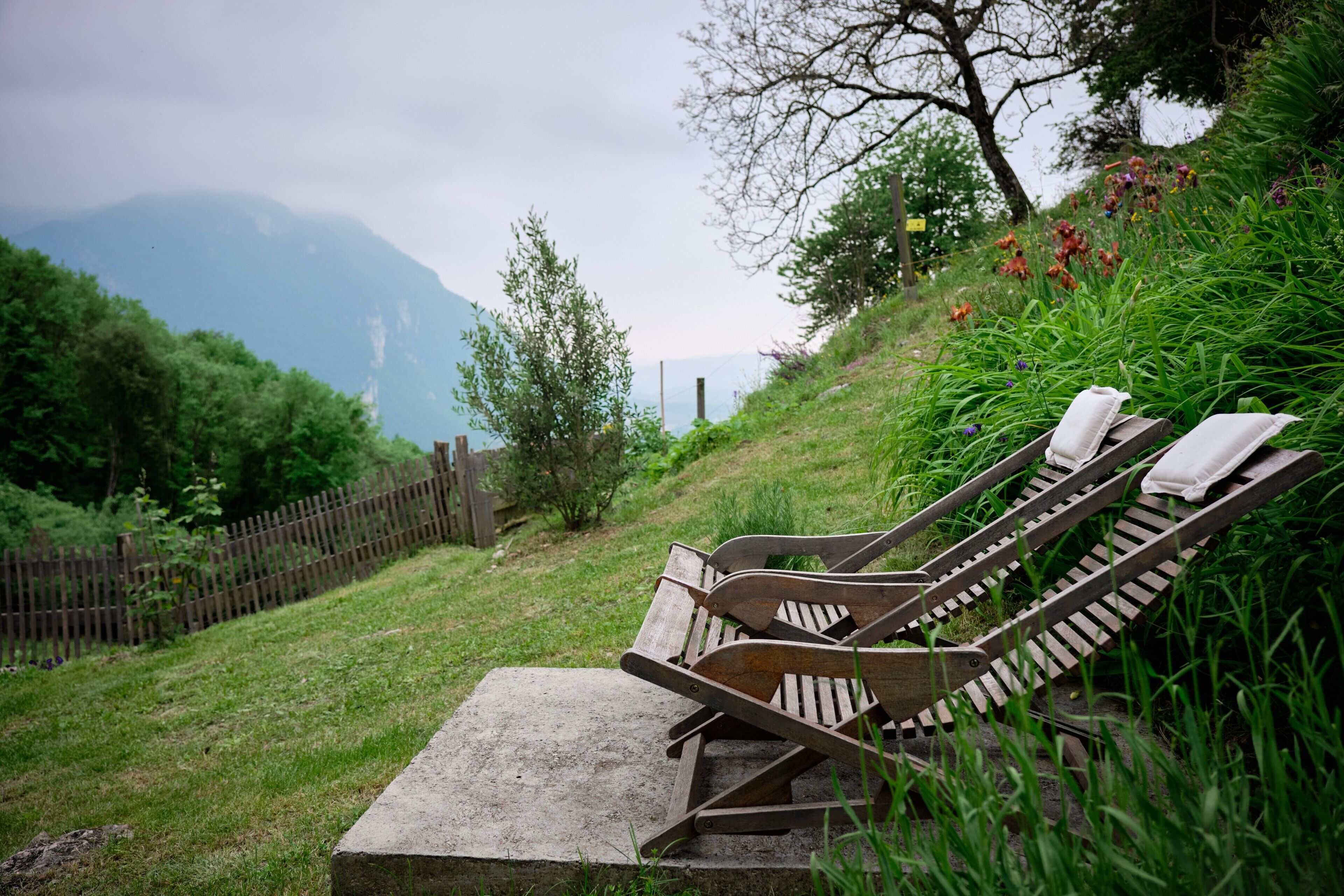 Two wooden lounge chairs with towels, set on a concrete platform, surrounded by greenery and a wooden fence, overlooking a mountainous landscape.
