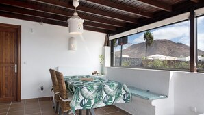 A dining area with a table covered in a tropical leaf tablecloth, wicker chairs, a hanging light fixture, and a view of mountains through a window. - Villa "Montaña Roja" with Private Pool, Terraces, and Wi-Fi (Playa Blanca)