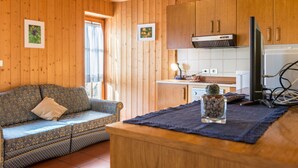 Een houten interieur met een bank, een kleine tafel met een plant en een keukenhoek met kasten en een koelkast.
