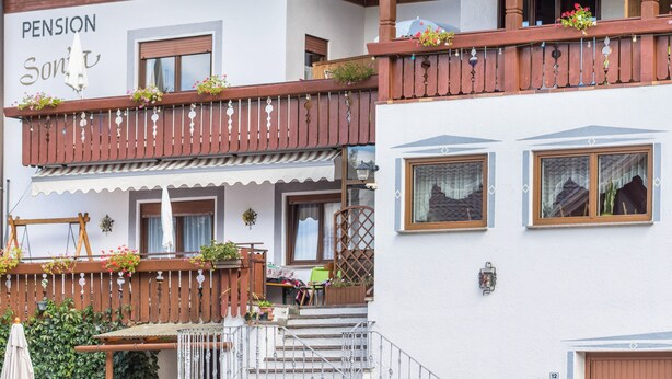 Een gebouw met een bord waarop 'PENSION Sonnt' staat, balkons met houten leuningen en bloembakken, een trap en een klein terras.