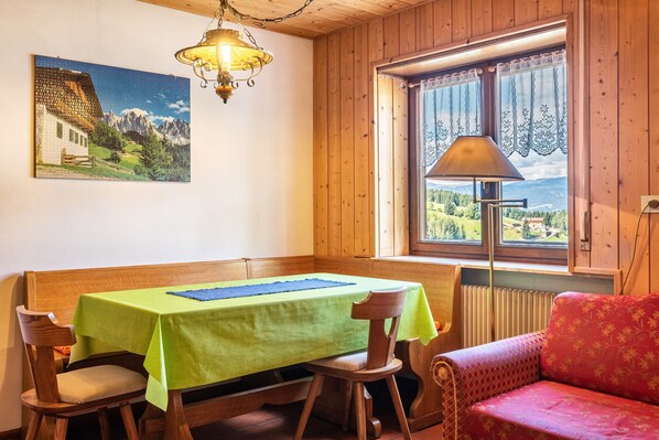 A dining area with a table, chairs, a bench, a sofa, a lamp, a window with curtains, and a painting on the wall.
