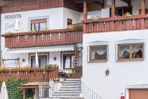 A building with a sign reading 'PENSION Sonnt.' balconies with wooden railings and flower boxes, a staircase leading to the entrance, and a small outdoor seating area with a table and chairs.