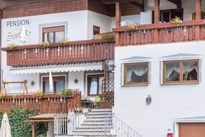 A building with a sign reading 'PENSION Sonnt.' balconies with wooden railings and flower boxes, a staircase leading to the entrance, and a small outdoor seating area with a table and chairs.