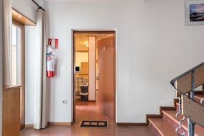 Un couloir avec un escalier en bois, un extincteur et une porte donnant sur une autre pièce.