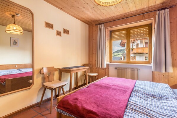 Una habitación de madera con una cama, un escritorio, una silla, un espejo y una ventana que da a vistas de edificios.