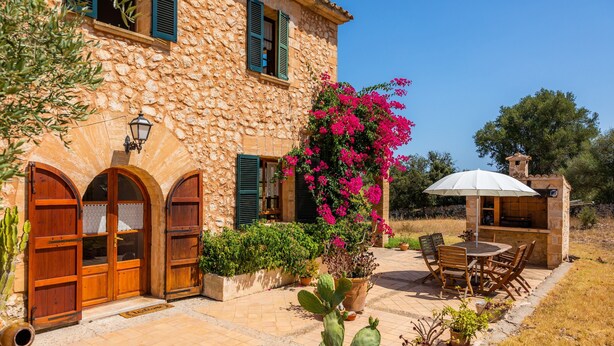 Ein Steinhaus mit Holztüren und -läden, eine Terrasse mit Tisch und Stühlen und ein Blumentopf mit rosa Blüten.