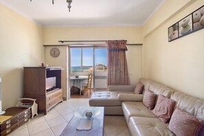 A living room with a sofa, a television, a wooden cabinet, a dining table, and a view through a glass door.