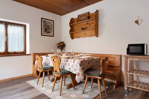 Zona de menjador amb taula de fusta preparada per a quatre, cadires amb coixins estampats, prestatgeria de fusta amb objectes decoratius i un televisor sobre un moble.