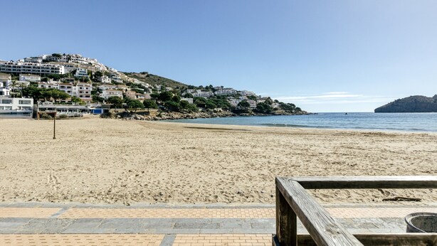 A beach with a wooden walkway leading to a building with multiple floors, situated on a hillside overlooking the sea. - Holiday Home "Casa en urb Cala Montgó" with Private Terrace and Wi-Fi (L'Escala)