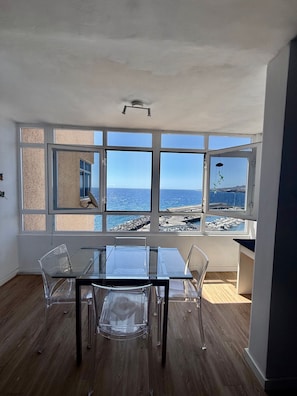 A room with a large window overlooking the sea, a glass table, and clear chairs.